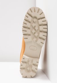 Botte en suede beige avec une semelle en caoutchouc blanc épaisse et texturée présentant un motif à clous pour une meilleure adhérence. La botte est partiellement appuyée contre une surface blanche.