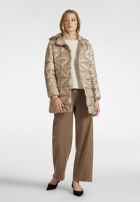 Vrouw in een beige gewatteerde pufferjack met een crèmekleurige sweater, bruine wijde broek en zwarte puntige slingback-schoenen, staand op een witte achtergrond.