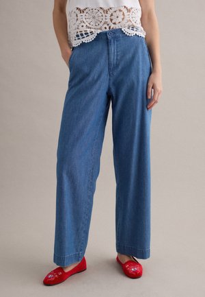 Donna che indossa jeans blu a gamba larga, top bianco in pizzo crochet e scarpe basse rosse ricamate, in piedi con le mani nelle tasche.