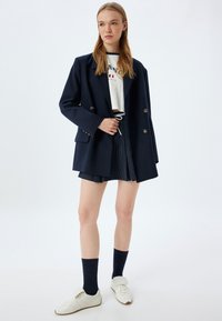 Marineblauwe dubbelgeknoopte blazer, witte cropped graphic tee, gestreepte shorts, witte sneakers en marineblauwe sokken. Schone, gestructureerde lijnen.