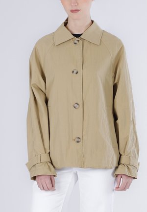 Beżowa kurtka na guziki z szerokim kołnierzem i regulowanymi paskami na mankietach, noszona przez osobę ubraną w białe spodnie i czarną bluzkę.