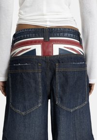 Pogled od zadaj na osebo, ki nosi temno modre kavbojke in pas z rdeče-belo-modrim motivom britanske zastave Union Jack, združene z belo majico z dolgimi rokavi.