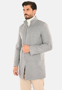 Cappotto in lana grigio chiaro con collo alto, tasche frontali e design pulito e lineare. Texture liscia, taglio a mezza lunghezza, vestibilità sartoriale.