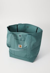 Teal tote bag med en struktureret design, lavet af holdbart stof, med to håndtag og et logo patch på forsiden. Indersiden er rummelig.