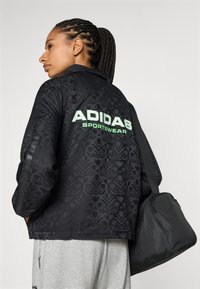 Svart sportjacka med präglat blommönster, grön "ADIDAS SPORTSWEAR" text på ryggen, i kombination med grå sweatpants och en svart väska.