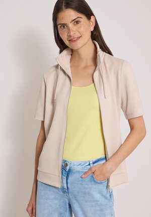 Jeune femme aux cheveux bruns portant une veste beige zippée à manches courtes, un haut jaune et un jean bleu clair, debout avec une main dans la poche.