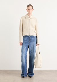 Beige tekstuur jack met rits en kraag, gecombineerd met blauwe wijde spijkerbroek en een lichte tote bag. Gesloten zwarte schoenen.