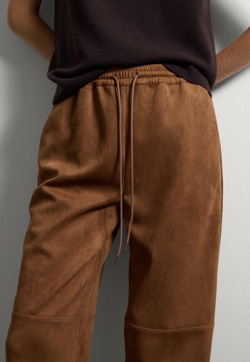 Joggers en suède brun avec une finition texturée, dotés d'une taille élastique et d'un cordon de serrage. Associés à un haut en tricot sombre.