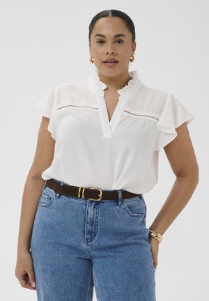 Kobieta ubrana w białą bluzkę z falbankami i koronkowymi detalami, niebieskie dżinsy, brązowy pasek, złote kolczyki koła oraz złota bransoletka, stojąca na jednolitym tle.