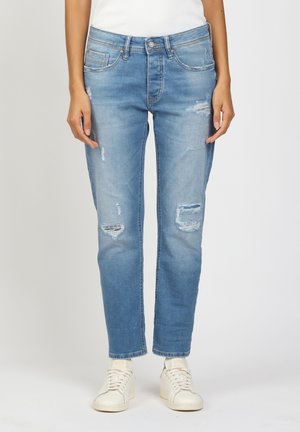 Person wearing light blue distressed jeans and white sneakers, standing against a plain white background.