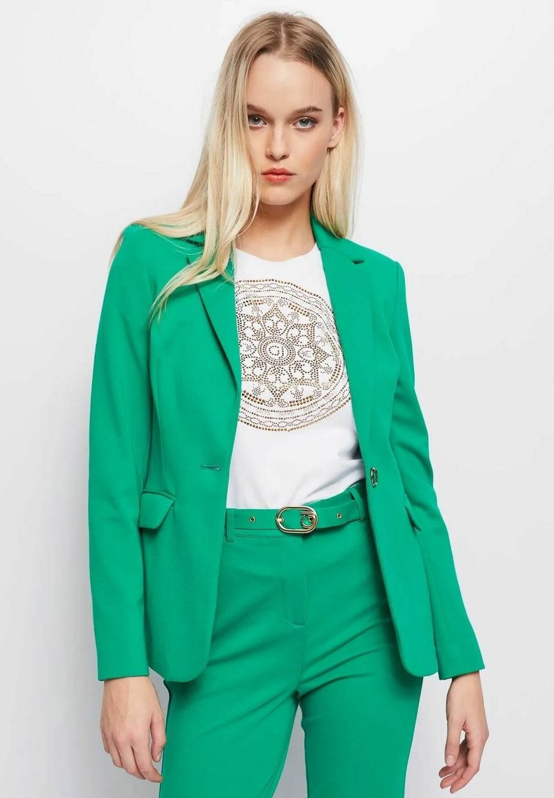 Groene blazer van gladde stof met notch-lapels en schoudervullingen, gecombineerd met een wit shirt met een gouden cirkelpatroon.