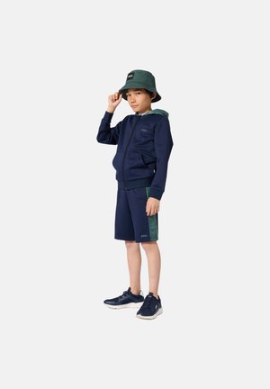 Marineblauwe hoodie met rits en groene accenten, bijpassende marineblauwe korte broek en donkere schoenen. Een groene bucket hat met een logo maakt de outfit compleet.