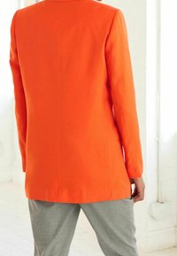 Giacca arancione brillante con un design sartoriale, maniche lunghe e taglio dritto. Indossata sopra pantaloni a scacchi bianco e nero.