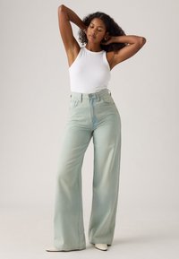 High-waisted, wide-leg jeans in light green denim, paired with a fitted white tank top. Smooth texture and classic five-pocket design.