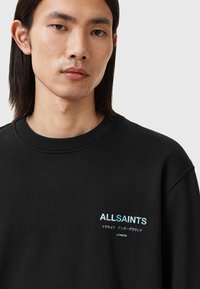 Sweatshirt noir à col rond, comportant un graphique sur le devant avec le texte "ALLSAINTS" en bleu et des textes supplémentaires en japonais.