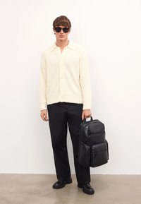 Krämfärgad texturerad cardigan, svarta byxor och svart ryggsäck. Solglasögon bärs. Ryggsäcken har flera fickor och en logotyp på framsidan.