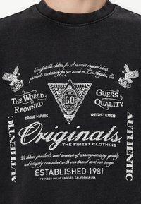 Sort sweatshirt med hvidt trykt tekst og grafik, herunder geometriske former og ørnerelaterede motiver. Ribbet crew neck og blød tekstur.