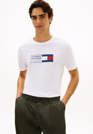 Mann iført en hvit Tommy Hilfiger t-skjorte med logo og grønne bukser, står med hendene i lommene mot en hvit bakgrunn.