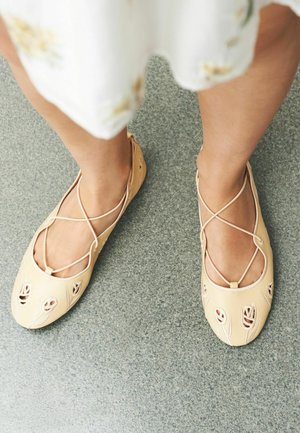 Nohy v béžových baletních balerinách s květinovými výřezy a zkříženými pásky, stojící na šedé texturované podlaze pod světle květinovými šaty.