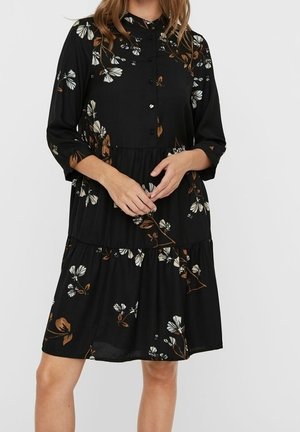 Zwarte knielange jurk met driekwartmouwen, knopen aan de voorkant en een witbruine bloemenprint, gedragen door een persoon die staat met handen zachtjes in elkaar gevouwen.