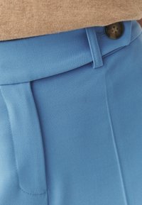 Pantalon en tissu bleu avec une finition texturée, doté d'un seul bouton foncé et d'une ceinture cintrée. Vue en gros plan du vêtement.
