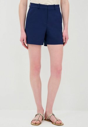 Shorts ajustés bleu marine en tissu lisse, avec une taille plate, des passants de ceinture et des poches latérales. Portés avec des sandales beiges.