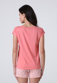 Top corallo a maniche corte realizzato in cotone morbido, caratterizzato da un fit rilassato e un collo a barca, abbinato a pantaloni shorts leggeri di colore rosa chiaro con fantasia.