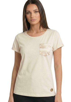 Une femme porte un T-shirt crème aux manches retroussées, avec une poche poitrine en satin et un petit logo, sur fond blanc.