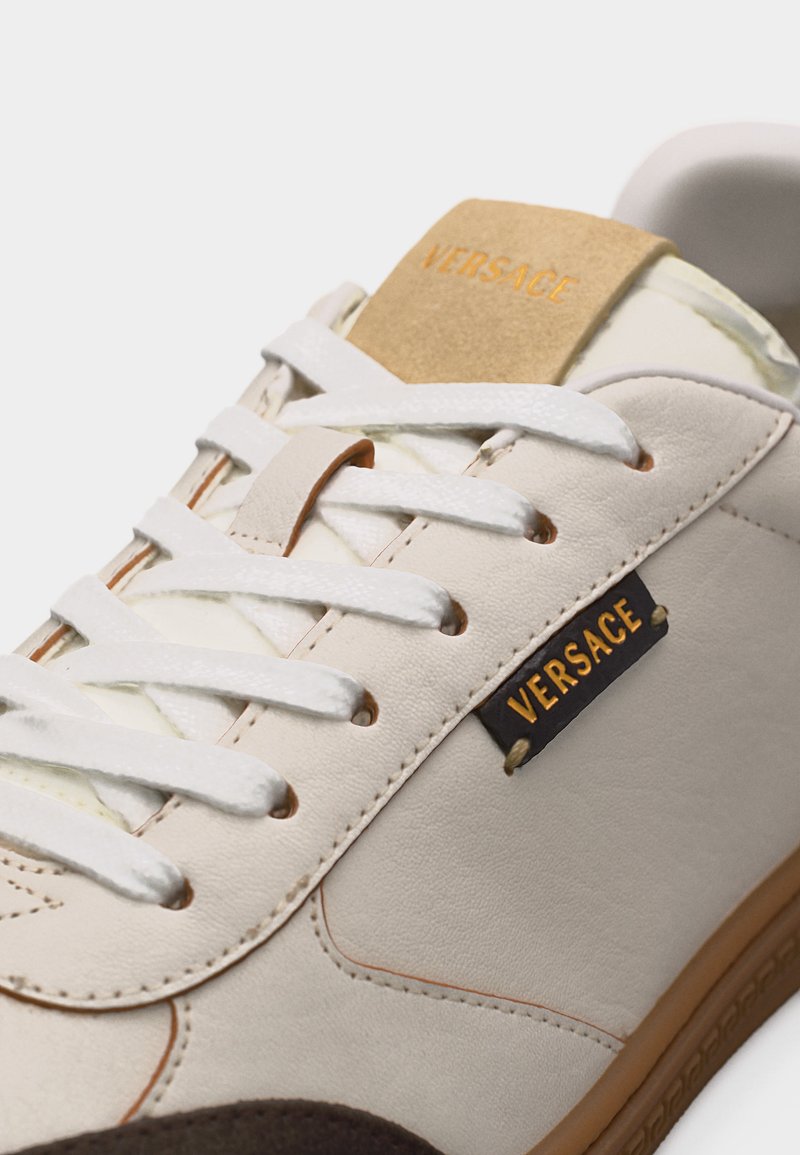 Primo piano di una sneaker in pelle color crema con lacci bianchi, suola marrone e logo "Versace" sulla linguetta e sull'etichetta laterale.