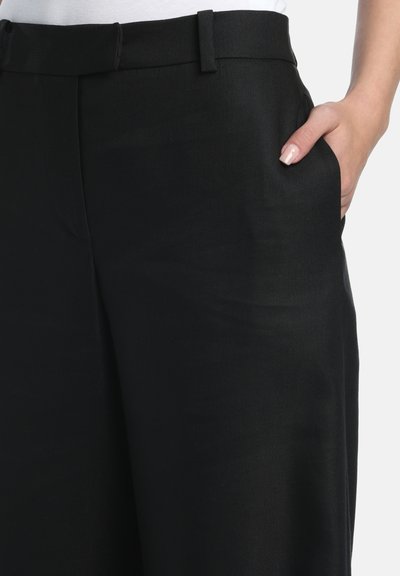 Gros plan sur un pantalon noir sur mesure avec passants pour ceinture et une main reposant dans la poche avant droite sur fond blanc.