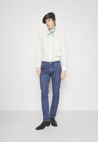 Camisa de lino blanca, bufanda azul, jeans ajustados azul oscuro y botas de cuero negras hasta el tobillo, complementadas con un sombrero beret negro.