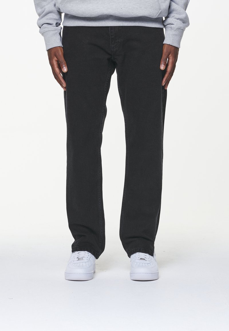 Svarta jeans av denim med rak passform, med minimal sömnad och två sidofickor. Matchat med vita sneakers mot en neutral bakgrund.