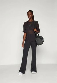 Μαύρο μπλουζάκι με δίχτυ και λογότυπο, συνδυασμένο με μαύρο κλασικό παντελόνι. Το μοντέλο κρατά μια μαύρη αθλητική τσάντα. Τα παπούτσια είναι λευκά. Απλή, αθλητική εμφάνιση.