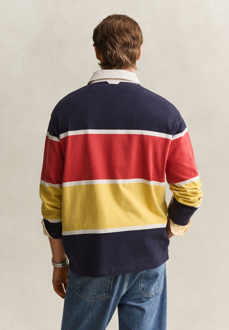 Mand iført langærmet rugbytrøje med marineblå, røde og gule vandrette striber, lys krave og blå jeans, som vender væk fra beskueren.
