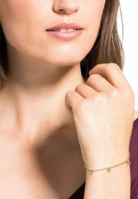 Primer plano de la parte inferior de la cara y el cuello de una mujer que lleva una delicada pulsera de oro con piedras verdes en su muñeca.