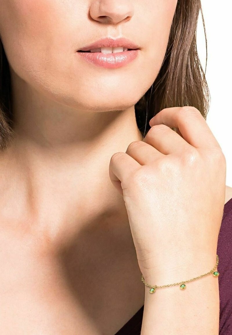 Primer plano de la parte inferior de la cara y el cuello de una mujer que lleva una delicada pulsera de oro con piedras verdes en su muñeca.