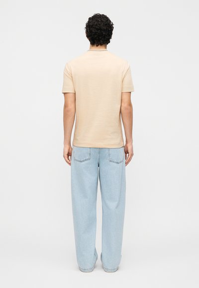 Άνδρας με σγουρά σκούρα μαλλιά, φορώντας μπεζ κοντομάνικο πουκάμισο και ανοιχτόχρωμο μπλε τζιν με φαρδιά παντελόνια, στέκεται με την πλάτη γυρισμένη σε λευκό φόντο.