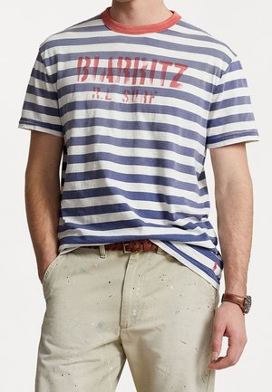 Pasiasta koszulka w kolorze granatowym i białym, z czerwonym ściągaczem przy szyi oraz graficznym napisem. Krótkie rękawy, luźny krój i miękki materiał bawełniany.