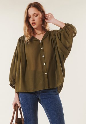 Femme portant un chemisier ample vert olive à boutons avec des manches volumineuses, jean bleu, collier chaîne en or, tenant un sac à main tressé marron.