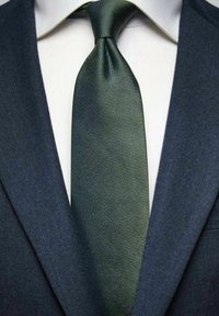 Tummansininen puku takki ja teksturoitu vihreä solmio. Solmiossa on hienovarainen kuvio, kun taas sen alla oleva paita on vaaleanvärinen.