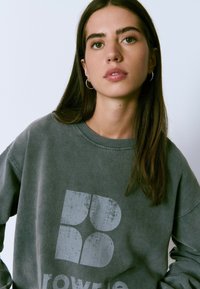 Sudadera gris con escote redondo y estampado de logo desgastado en un gris más claro. La tela parece suave con un acabado ligeramente texturizado.