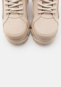 Par beige lædersneakers med tykke snørebånd og tekstureret sål set ovenfra på en hvid baggrund.