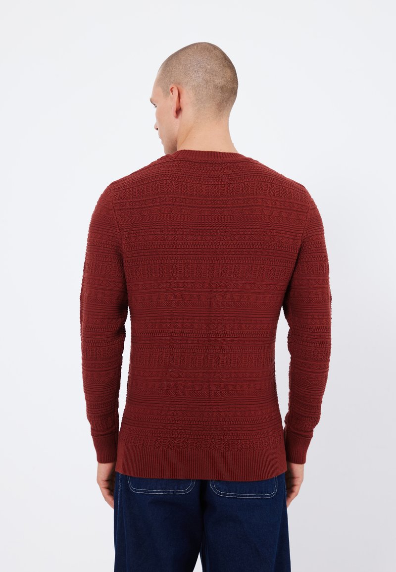 Maglione rosso a fantasia con maniche lunghe, che presenta un design a maglia testurizzata e polsini a coste, abbinato a pantaloni in denim blu.