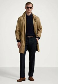 Casaco tipo trench bege sobre um suéter azul-marinho, combinado com calças azul-marinho com riscas. Sapatos de couro castanho e óculos de sol completam o visual.