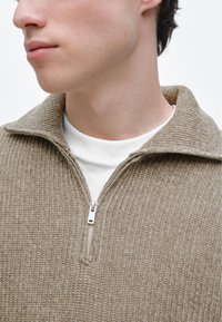Pull beige à fermeture éclair avec un design côtelé texturé, doté d'un col montant et d'une fermeture éclair argentée, porté sur un t-shirt blanc à col rond.