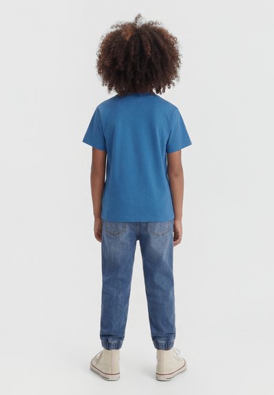 Enfant aux cheveux bouclés portant une chemise bleue à manches courtes, un pantalon jogger en denim et des baskets montantes de couleur claire, debout de dos.