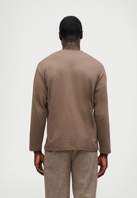 Muškarac okrenut leđima u dugom smešnom smešnom smeškom bež džempera i bež teksturiranim hlačama pred jednostavnom belom pozadinom.