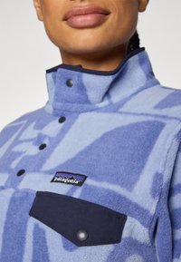 Fleece-Pullover in Hellblau mit dunkelblauen abstrakten Mustern, hohem Kragen, Druckknöpfen und einer schwarzen Brusttasche mit einem Logo.