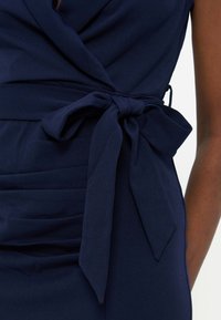 Φόρεμα σε ναυτικό μπλε χρώμα με ζώνη από ύφασμα δεμένη σε φιόγκο στη μέση, φορεμένο από άτομο με σκούρο χρώμα δέρματος σε λευκό φόντο.