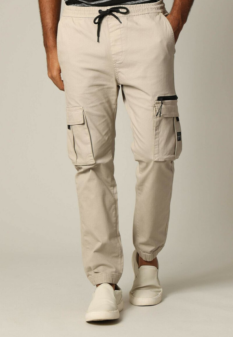 Beige Cargohosen mit elastischen Bündchen, mehreren Taschen und einer Kordelzug-Taille. Hergestellt aus leichtem Stoff mit schwarzen Reißverschlüssen.